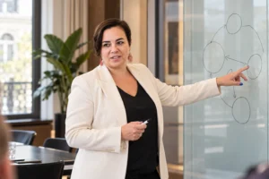 An Vermeulen in een ivoorkleurige blazer wijst naar een eenvoudig schema op een glazen bord.