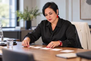 An Vermeulen in een zwarte blouse ordent blanco kaarten op een houten tafel.
