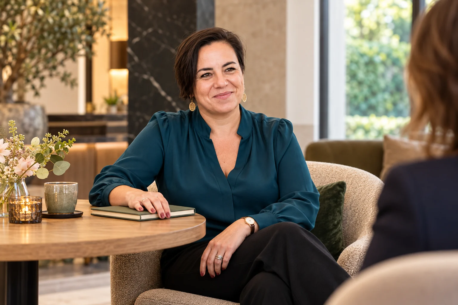 An Vermeulen in een petrolkleurige blouse glimlacht naar een gesprekspartner met haar hand op een gesloten notitieboek.