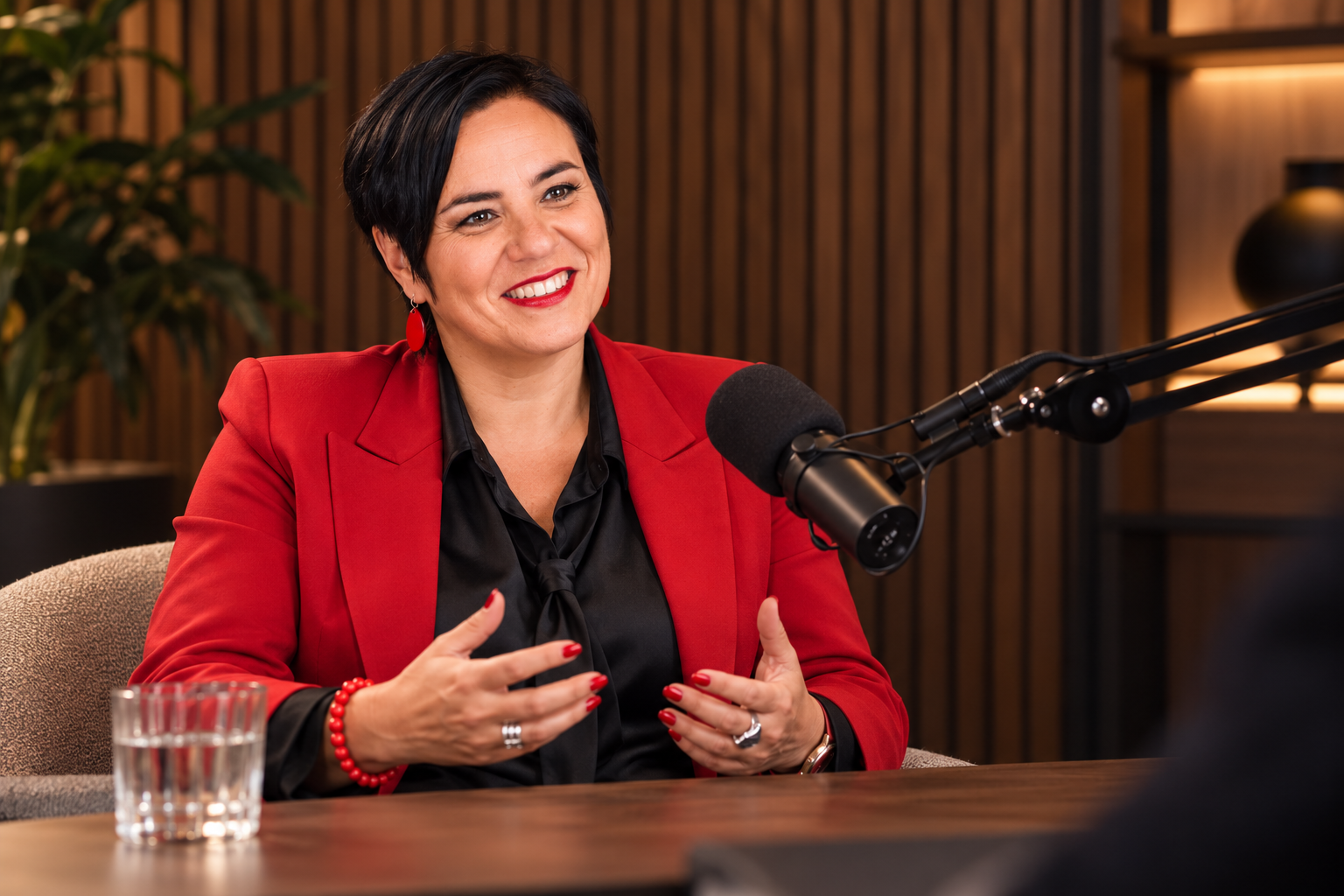 An Vermeulen in een rode blazer spreekt aan een tafel met een podcastmicrofoon.